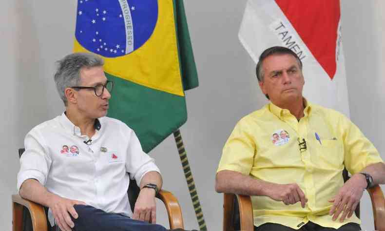Romeu Zema e Jair Bolsonaro sentados lado a lado com as bandeiras do Brasil e Minas Gerais ao fundo