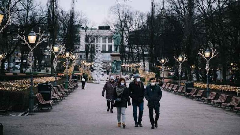 Fotografia colorida mostra jovens de mscara caminhando em uma boulevard coberta por luzinhas de Natal