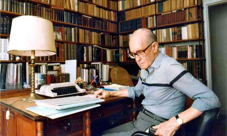 Carlos Drummond sentado na escrivaninha de sua biblioteca, com estantes cheias de livros ao fundo 