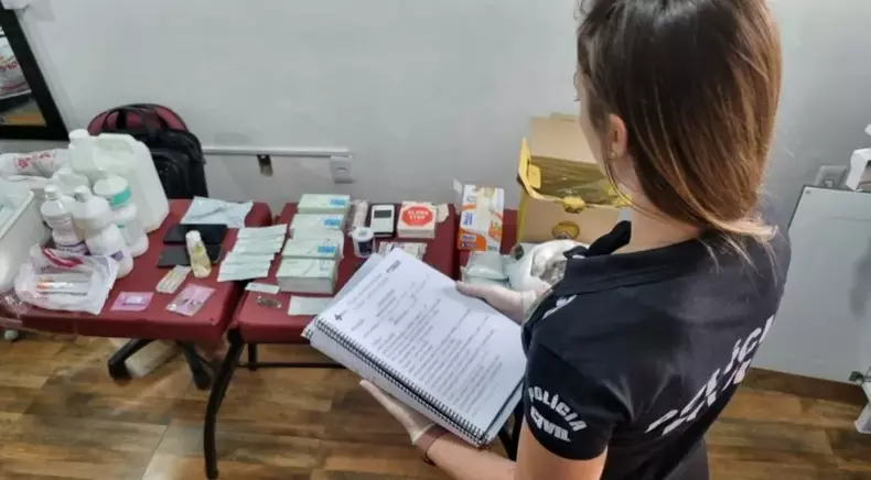 Mulher presa em Goiânia fazia cirurgias plásticas desde 2021(foto: CNN Brasil/Reprodução) Agente da Polícia Civil fiscalizando/olhando para mesa com produtos apreendidos da cirurgiã