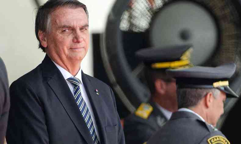 A imagem mostra o presidente Jair Bolsonaro olhando de lado. Ele  um homem branco de cabelo preto, ele est usando terno e uma gravata listrada. Atrs dele alguns militares esto de costas