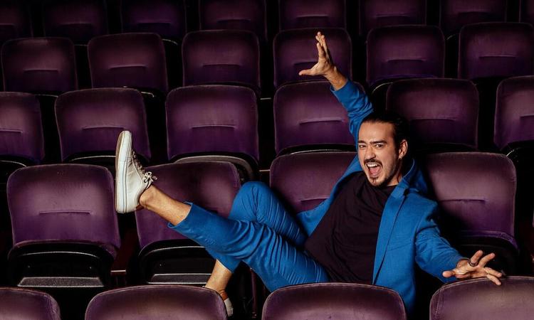 Humorista Marcos Veras, sorrindo, est deitado em cadeiras de plateia de teatro
