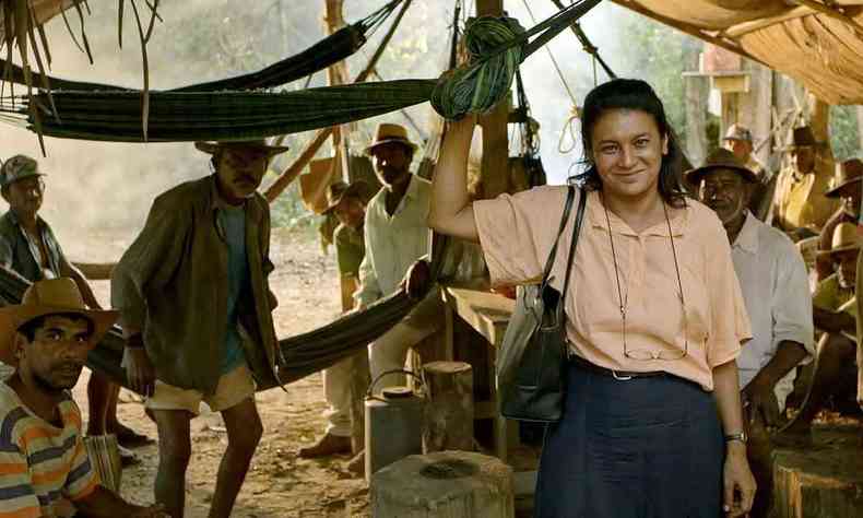 Vestida com blusa clara e saia preta, em acampamento cheio de trabalhadores, a atriz Dira Paes sorri, em cena do filme Pureza