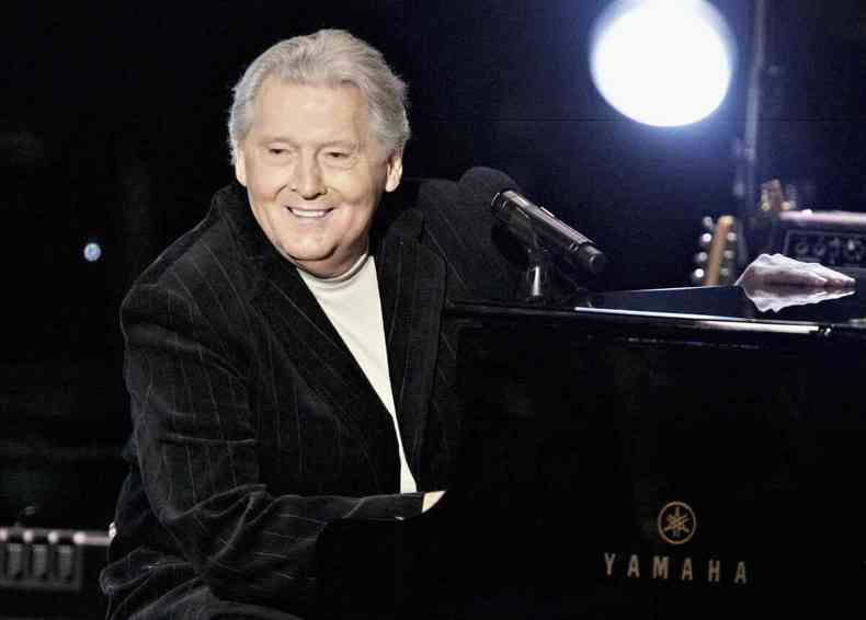 Cantor e pianista Jerry Lee Lewis de blazer preto e blusa branca sentado em frente a um piano