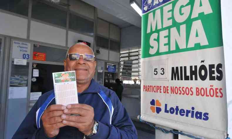 Ao lado de um cartaz da Mega-Sena de 53 milhes de reais, apostador sorri e mostra os volantes 