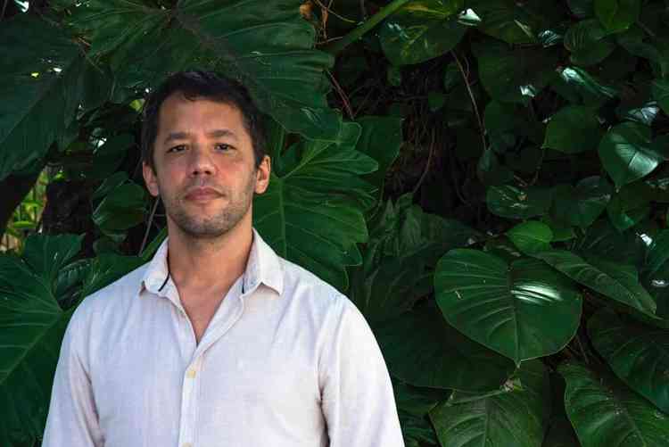 Na foto, o escritor baiano Itamar Vieira Junior de camisa branca, em frente a uma folhagem, olha para a cmera