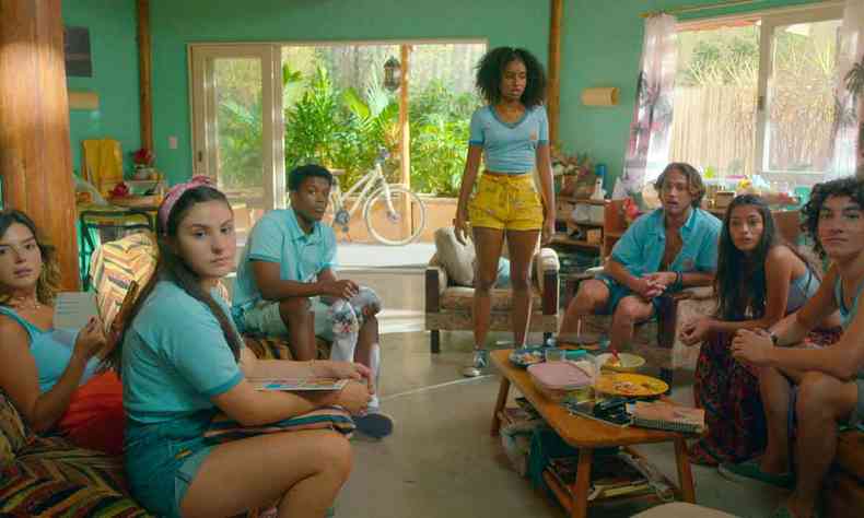 Sete jovens atores da série Temporada de verão estão numa sala de restor, sentados no sofá, com roupas praianas