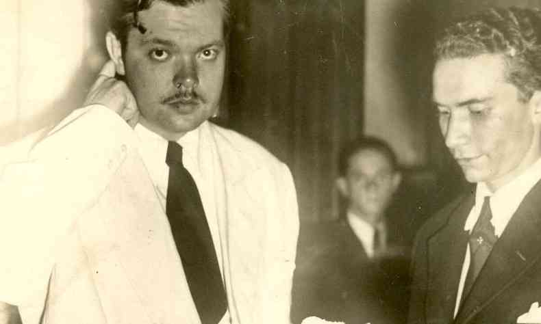Orson Welles olha para a cmera, vestindo terno branco, e  entrevistado por reprter do Estado de Minas em sua visita a BH, em 1942