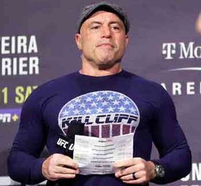 De bon e camisa de manga comprida, Joe Rogan segura papel com as duas mos 