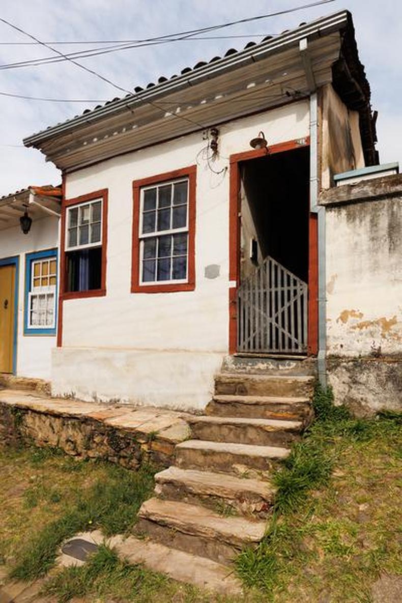 Casa antiga branca, com duas janelas e uma porta com moldura marrom; na entrada, uma escada com sete degraus