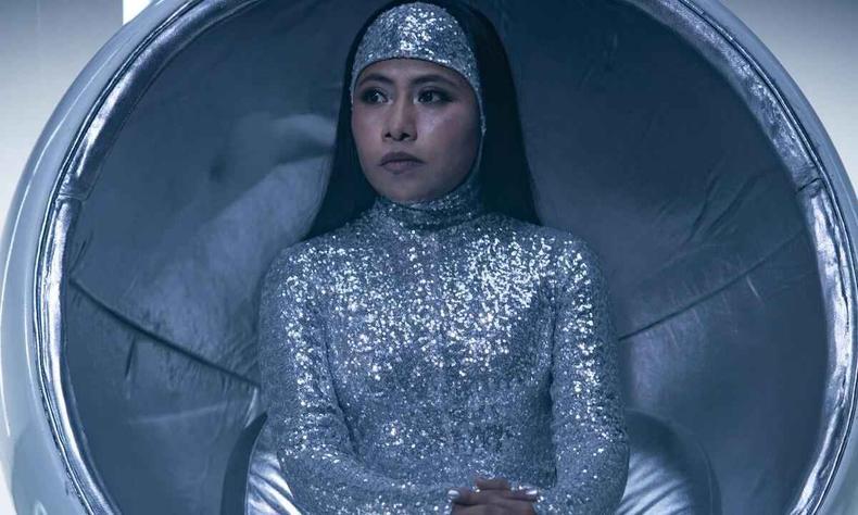 A atriz Yalitza Aparicio como Lua em srie, com roupa prateada, sentada em banco em formato de ovo
