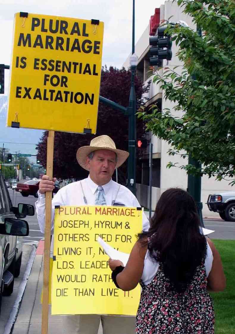Manifestante na rua segurando placa defendendo poligamia conversa com mulher, que aparece de costas