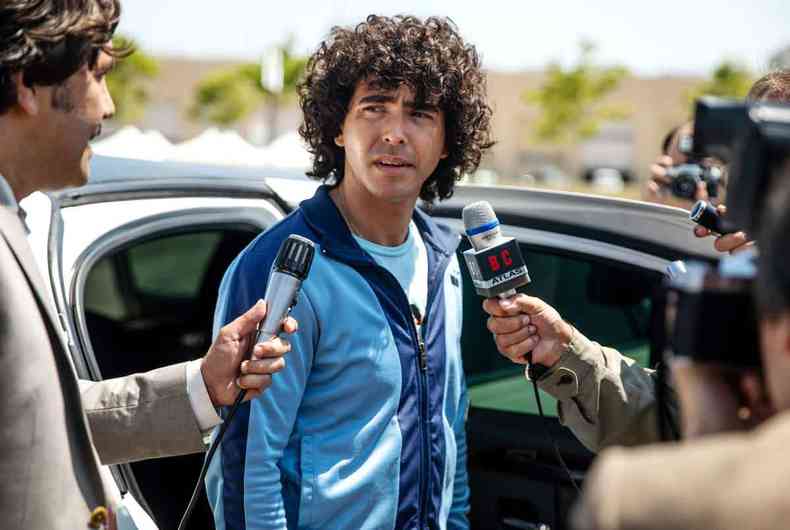 Ator que interpreta Maradona, de pé, vestido de moletom, é abordado por dois repórteres em cena de série