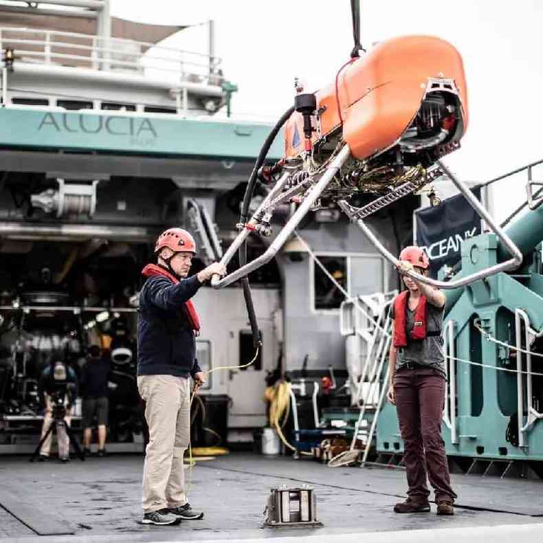 Veículo robótico subaquático Orpheus sendo manipulado por dois profissionais usando capacetes e coletes salva-vidas