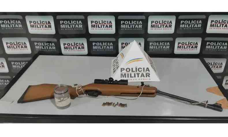 Carabina e munição em cima de uma mesa com banner da polícia civil ao fundo