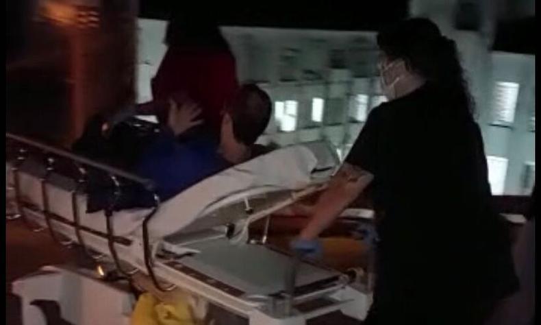 Homem branco est deitado em uma maca sendo levado por uma mulher de cabelos presos