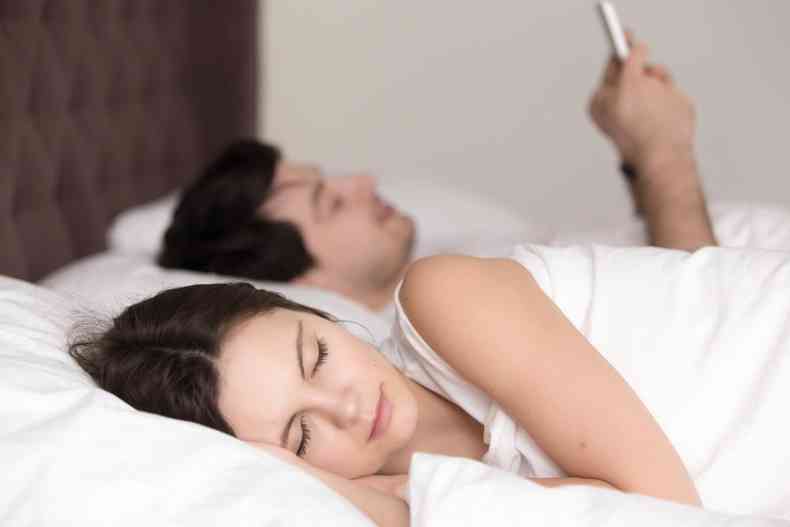 Jovem mulher dormindo enquanto seu namorado usa smartphone na cama