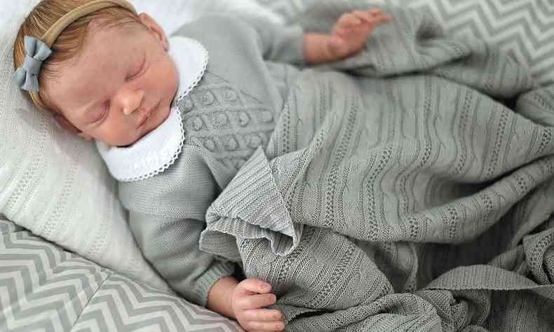 Boneco de beb sobre a cama, com os olhos fechados, usa roupinha cinza e lao na cabea