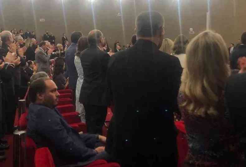 Carlos Bolsonaro sentado durante cerimnia de posse, enquanto outros presentes aplaudem de p