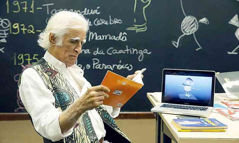 Vestindo camisa branca e colete, sentado em cadeira, com quadro negro atrs de si, Ziraldo l um livro 