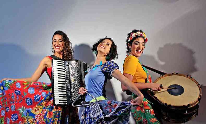 vestidas com roupas à moda caipira e segurando seus instrumentos, as integrantes do trio Mana Flor sorriem para a câmera, de pé, em ambiente neutro