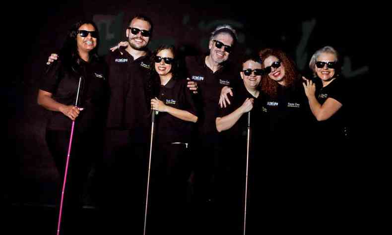 Sete atores da Companhia de Teatro Cego, lado a lado, sorriem para a câmera usando óculos escuros, roupas pretas e bengalas