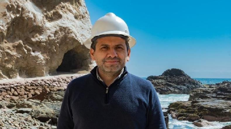 Gerardo Espindola Rojas posa para foto com capacete em frente a praia com caverna