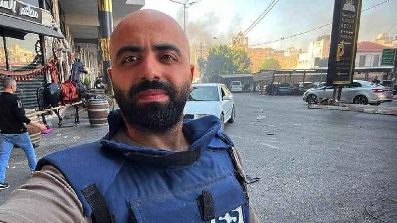 Alaa Daraghme com colete a prova de balas em rua, fazendo selfie, com fuma�a atr�s