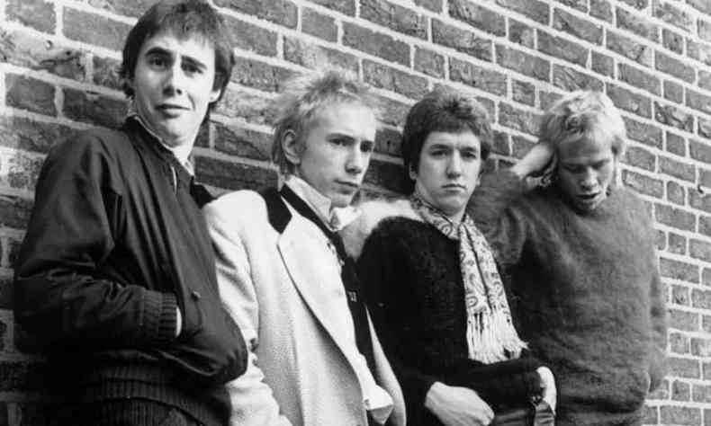 Encostados numa parede de tijolos aparentes, quatro integrantes da banda Sex Pistols olham para a cmera