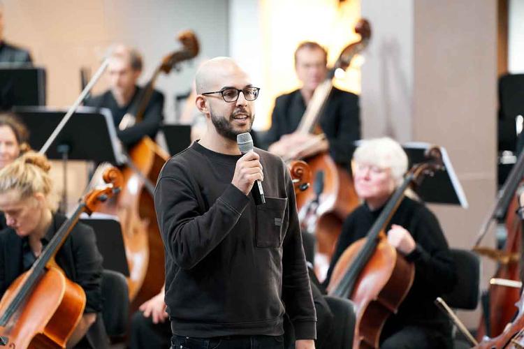 Leonardo Silva fala ao microfone, tendo msicos de orquestra atrs de si