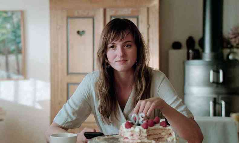 A atriz Renate Reinsve vestida de branco, sentada  mesa, com um bolo de aniversrio e velas marcando 30  sua frente, em cena de a pior pessoa do mundo