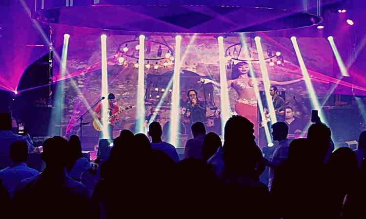 Cantor da banda U2 Cover Brasil faz show no palco, sob luzes de holofotes e luz cor de rosa. Ao fundo, telo mostra bailarina
