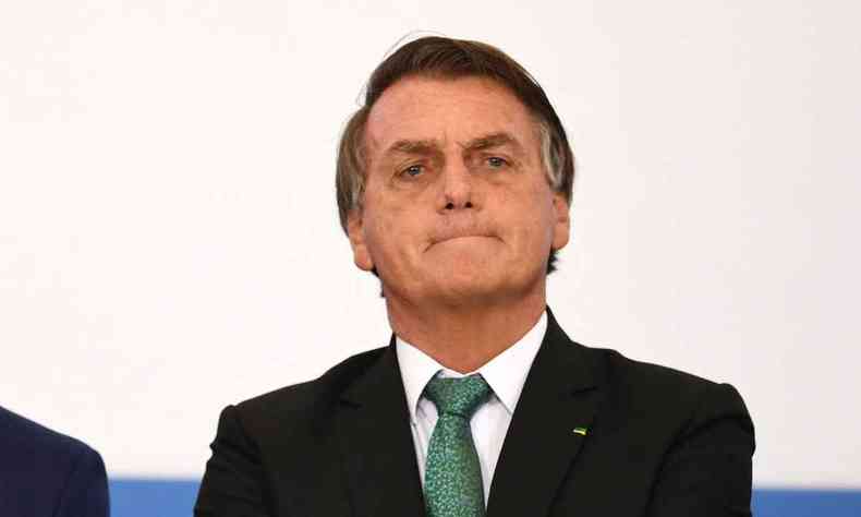 Bolsonaro de terno preto, camisa branca e gravata verde com expresso sria em um funco branco