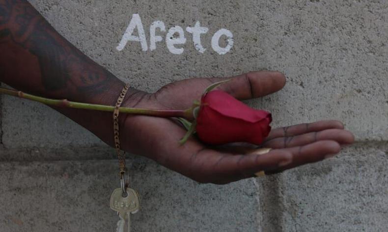 Uma mo negra segura uma rosa vermelha com delicadeza. Na parede de concreto ao fundo, l-se na parede 'Afeto'