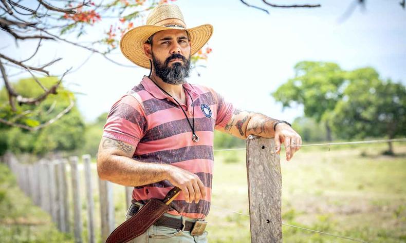 Ator Juliano Cazarr, da novela Pantanal, posa de blusa listrada, chapu e amparado em cerca 