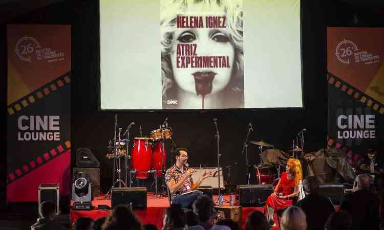 Atriz Helena Ignez est sentada no palco, durante homenagem prestada a ela na Mostra de Tiradentes. ao fundo, imenso banner traz foto da artista