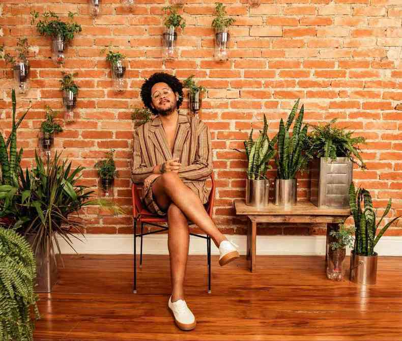 Rapper paulistano Emicida est de pernas cruzadas, ao lado de vasos de plantas, sentado em varanda com parede de tijolinhos