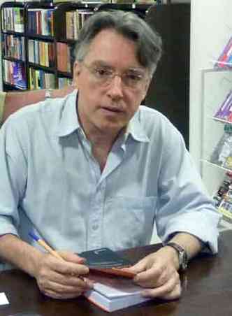 O escritor Bráulio Tavares, sentado, autografa um livro e olha para a câmera 