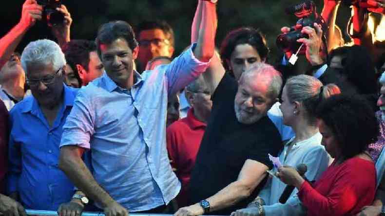 Haddad e Lula levantando os braos juntos, rodeados por outras pessoas em evento