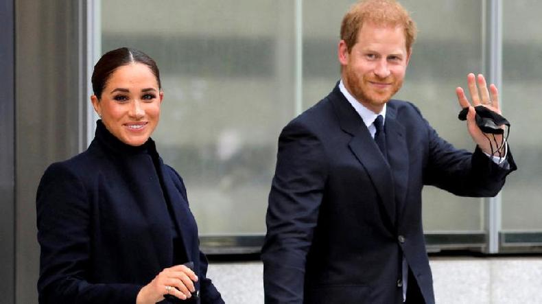 Meghan e Harry sorrindo para foto enquanto caminham com trajes formais em rea externa