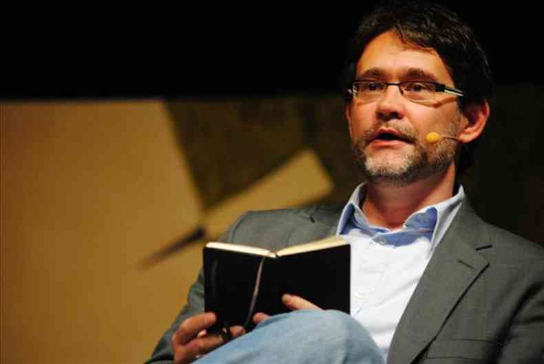 Professor e pesquisador João Cezar de Castro Rocha, de barba, óculos, camisa branca e blazer cinza, segura um livro
