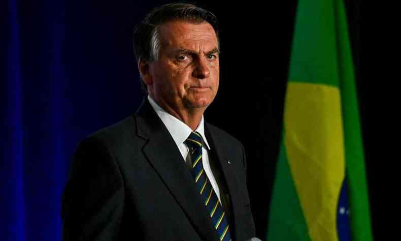 Ex-presidente Jair Bolsonaro. Ele  um homem branco, de cabelos grisalhos e usa terno. No fundo uma bandeira do Brasil