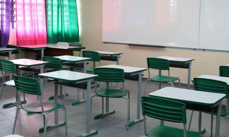 Sala de aula vazia, vista do fundo da sala, mostrando carteiras e quadro.  esquerda aparecem janelas e cortinas coloridas. 