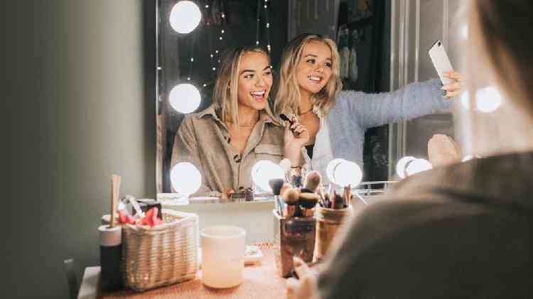 Duas jovens se maquiando e fazendo selfie em frente ao espelho