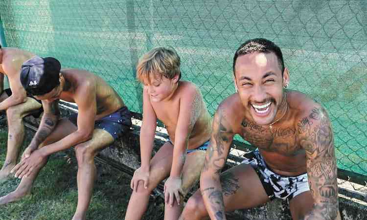 Neymar, sem camisa e de calo, sorri ao lado do filho Davi Lucca e de amigos no banco de quadra esportiva