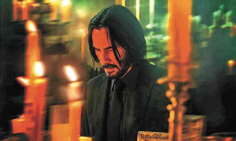 Vestido de preto em ambiente com velas, Keanu Reeves abaixa a cabea em cena de 'John Wick 4: Baba Yaga'
