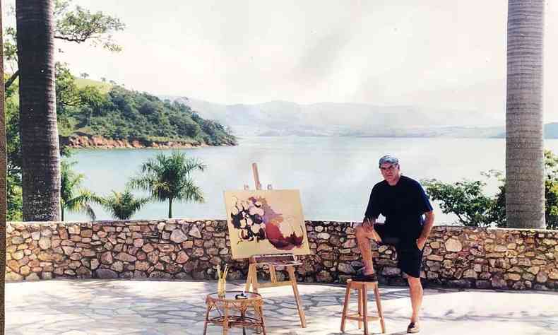 Pintor Fernando Palma ao lado de tela, no jardim. Ao fundo v-se a represa de Furnas