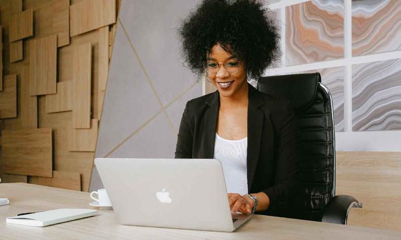 Mulher negra, com cabelo black power, usando blusa branca e blazer preto trabalha  mesa com um macbook 