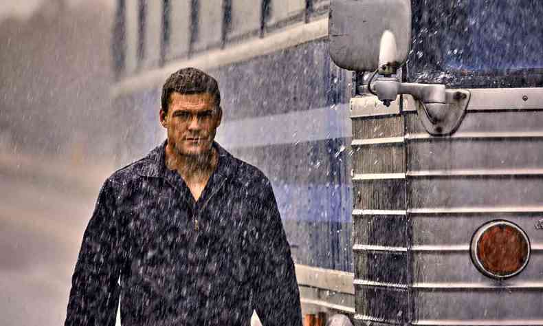 De casaco preto, Alan Ritchson (Jack Reacher) caminha na chuva, ao lado de um nibus