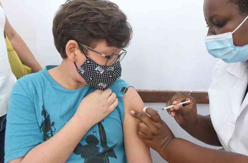 Menino de camisa azul e mscara recebe a vacina de uma enfermeira 